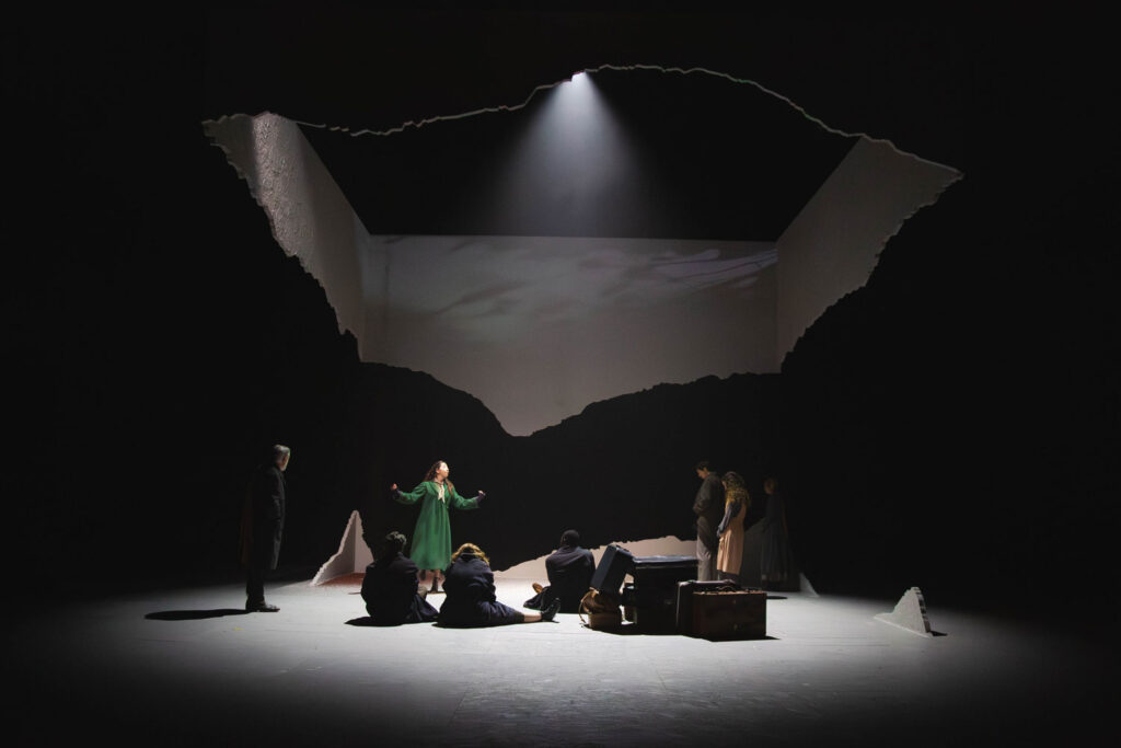 Una escena teatral dramática muestra a una mujer vestida de verde de pie, con los brazos en alto, rodeada de gente sentada o de pie. La escenografía presenta paredes desgarradas y dentadas, y un foco que ilumina desde arriba, resaltando el centro del escenario.