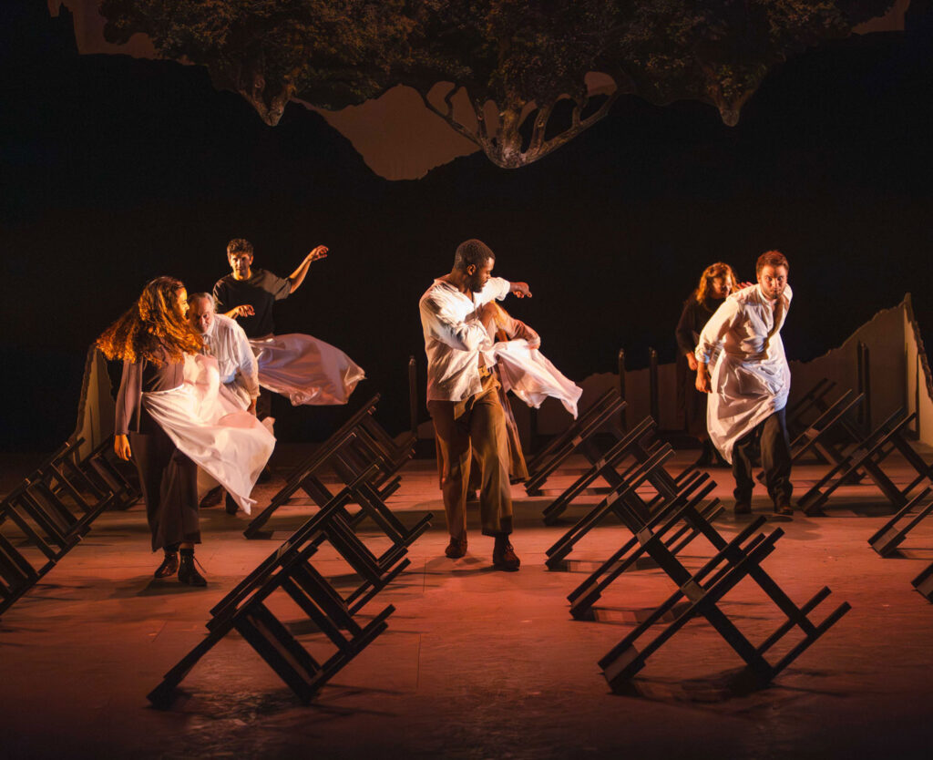 Seis bailarines, vestidos con camisas blancas y pantalones oscuros, bailan con energía entre filas de sillas volcadas en un escenario espectacular y con poca luz, bajo una estructura surrealista que parece un árbol flotante.