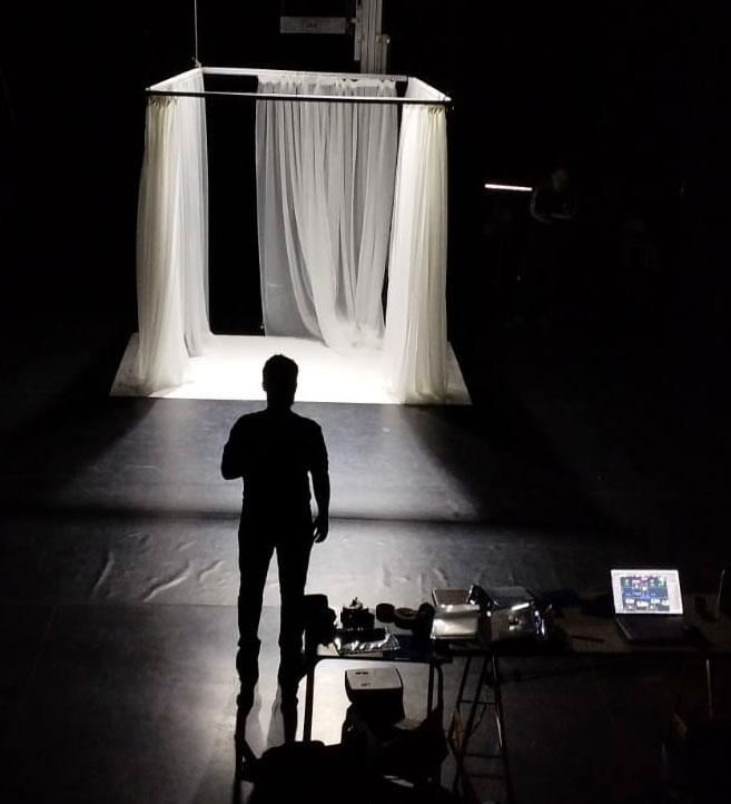 Se ve la silueta de una persona de pie junto a una mesa con material técnico, de cara a un pequeño escenario con cortinas blancas transparentes que cuelgan de un marco, todo ello iluminado en una sala que, por lo demás, está a oscuras. Kevin Dornan.