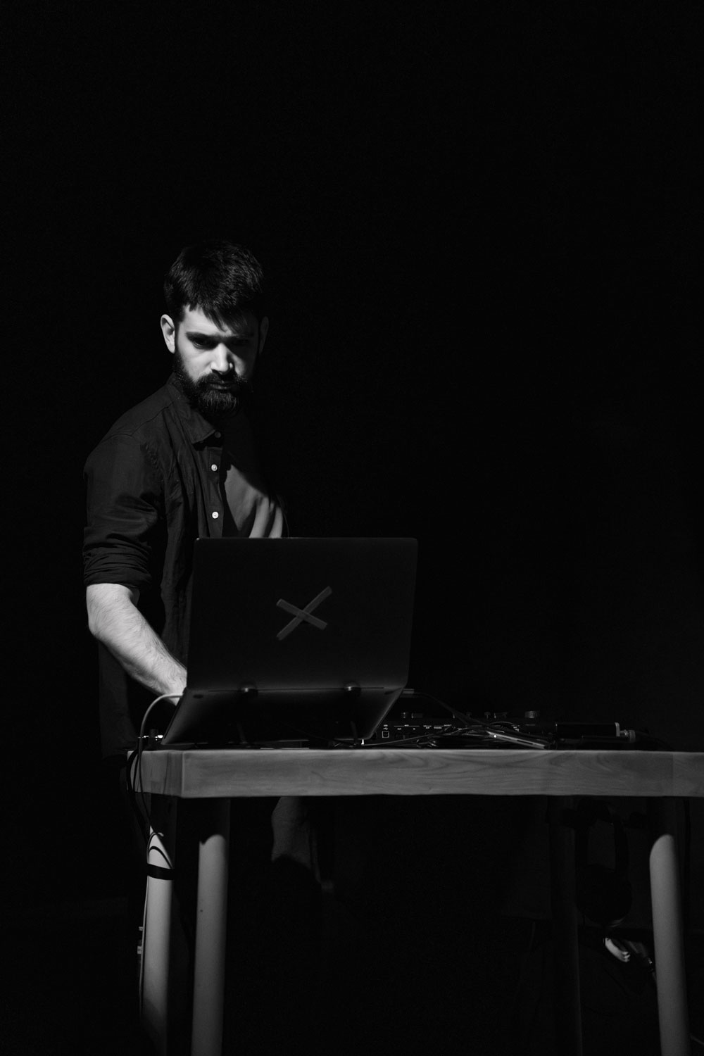 Kevin Dornan. Un hombre con barba está de pie junto a una mesa, utilizando un ordenador portátil y un equipo de DJ, iluminado por una única luz en una habitación que, por lo demás, está a oscuras.