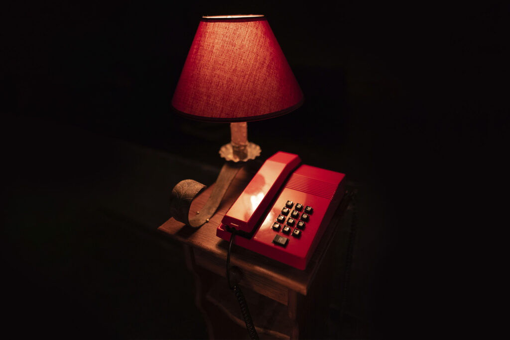 Un teléfono rojo de disco descansa sobre una pequeña mesa de madera junto a una lámpara con pantalla roja encendida, que proyecta un cálido resplandor en una habitación a oscuras.