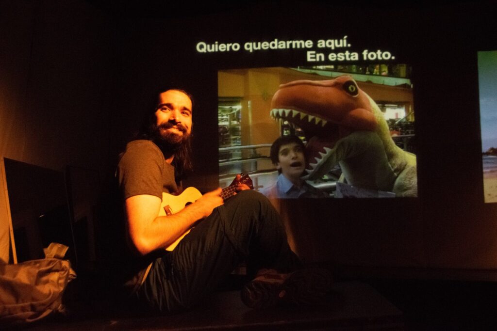 Una persona con barba está sentada en una luz tenue, sonriendo y sosteniendo una pequeña guitarra. Detrás de ella hay una proyección de un niño con una gran maqueta de dinosaurio y un texto en español: «Quiero quedarme aquí. En esta foto». El estilo de Kevin Dornan se refleja claramente en la escena.