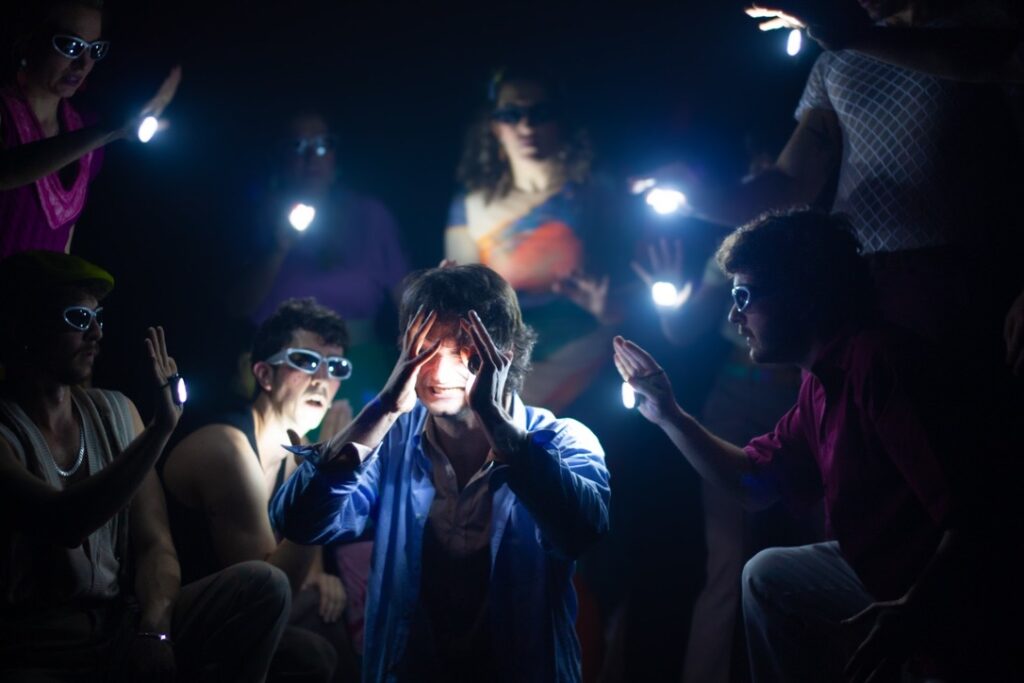 Una persona con una chaqueta azul está rodeada de otras que sostienen luces brillantes en las manos, lo que crea un efecto espectacular en la oscuridad. La figura central se tapa la cara mientras las demás se inclinan hacia ella.