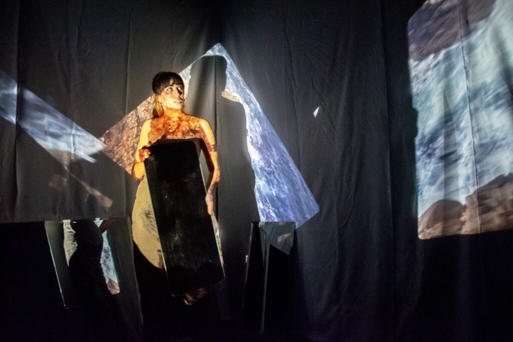 Kevin Dornan se encuentra en el escenario sosteniendo un gran objeto rectangular, iluminado por proyecciones dramáticas y abstractas de rocas o agua sobre un fondo oscuro, lo que crea una atmósfera surrealista y artística.