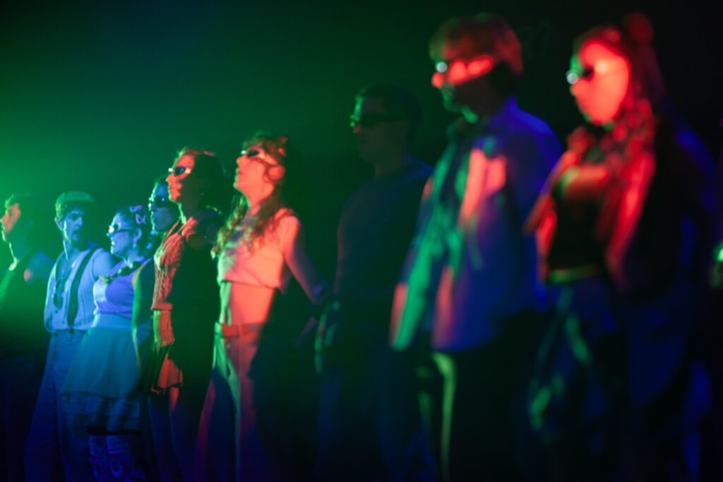 Un grupo de personas está de pie, una al lado de la otra, bajo una iluminación colorida y espectacular, con trajes y gafas de sol, mirando hacia delante en un escenario con poca luz.