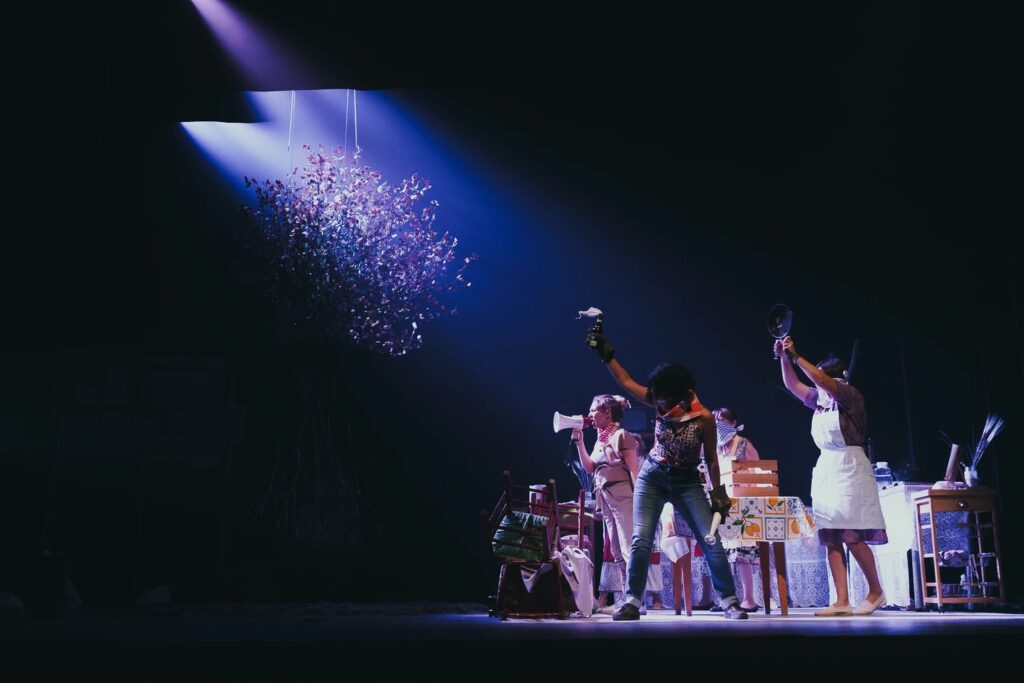 Un grupo de artistas en un escenario con poca luz, posando con energía bajo un foco, rodeados de atrezo y pupitres, con ramas que cuelgan por encima, lo que crea una atmósfera dramática y teatral.