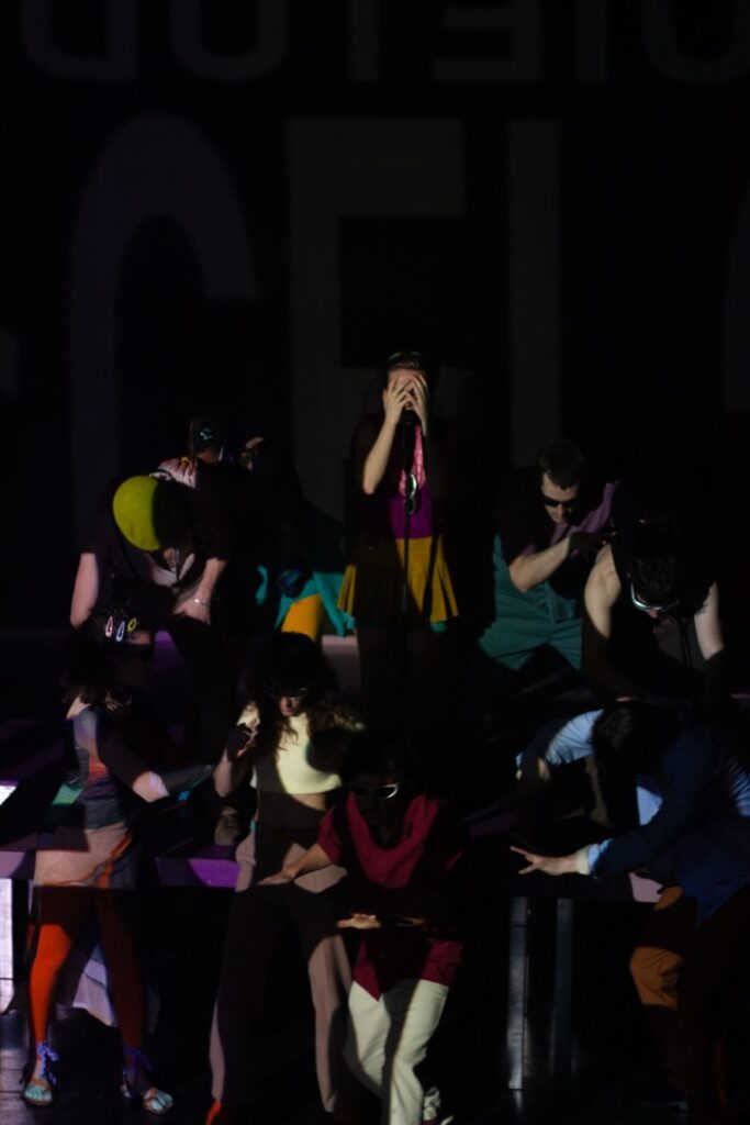 Un grupo de personas en el escenario lleva ropa de colores vivos y máscaras, y la iluminación teatral proyecta sombras. Una persona se encuentra de pie en el centro, con algo pegado a la cara, mientras que las demás están agachadas o hacen gestos en poses dinámicas.