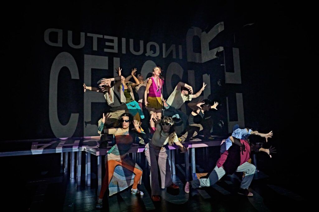 Un grupo de artistas con trajes de colores vivos adopta poses dramáticas en un escenario a oscuras, con sombras marcadas y un texto grande y reflejado proyectado en el telón de fondo que tienen detrás.