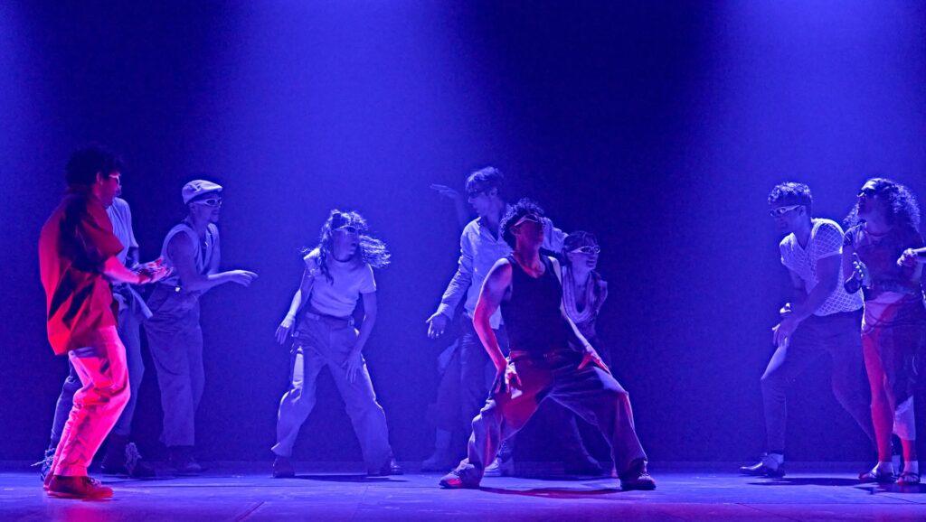 Un grupo de bailarines actúa con energía en el escenario bajo una espectacular iluminación púrpura, con poses expresivas y movimientos dinámicos que transmiten emoción e intensidad.