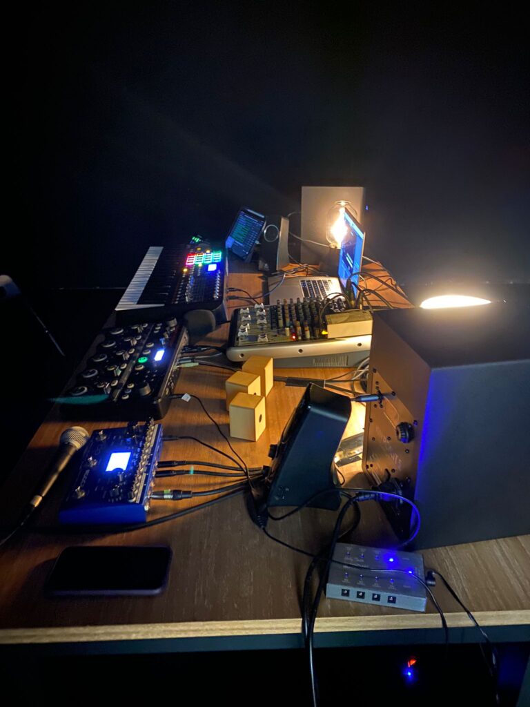Una mesa de estudio con poca luz, equipada con material de música electrónica —entre el que se incluyen un teclado, una mesa de mezclas, interfaces de audio, altavoces y un micrófono—, es decir, el equipo de Kevin Dornan, en la que los controladores y los cables están dispuestos de forma ordenada sobre una superficie de madera.