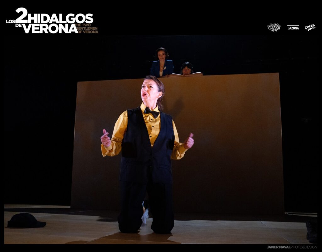Un actor con una camisa amarilla y un chaleco oscuro está arrodillado en el centro del escenario, gesticulando con ambas manos. Dos personas observan desde detrás de un telón de fondo alto y sencillo. La escena es dramática y está poco iluminada. El texto reza: «Los 2 Hidalgos de Verona».
