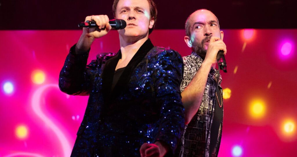 Dos hombres cantan juntos en el escenario, cada uno con un micrófono en la mano. Uno lleva una chaqueta azul brillante y el otro, una camisa estampada. El fondo está iluminado por luces de colores, desenfocadas, que crean un ambiente animado.