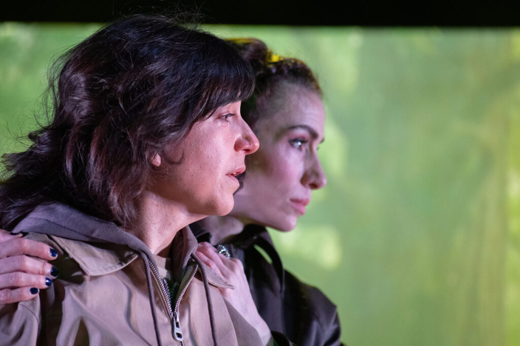 Dos mujeres están de pie muy juntas, mirando fijamente hacia delante. Una tiene la mano sobre el hombro de la otra. Ambas parecen serias, con un fondo verde difuminado detrás de ellas.