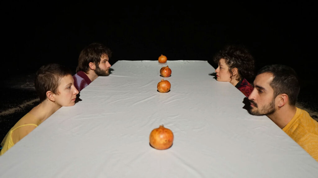 Cuatro personas se miran unas a otras desde los lados opuestos de una mesa cubierta con un mantel blanco, con tres granadas distribuidas a igual distancia en el centro. El fondo es oscuro, lo que hace que la atención se centre en la escena.
