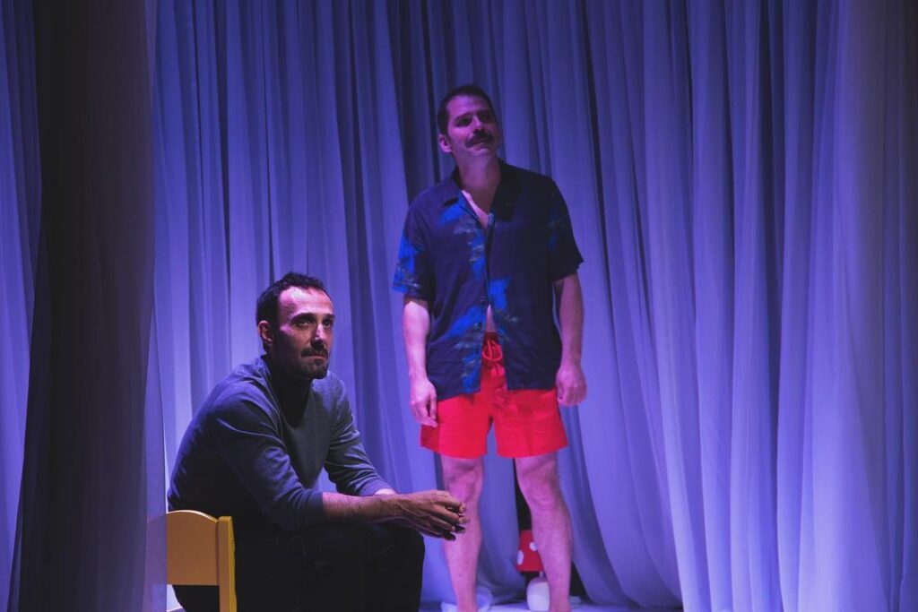 Hay dos hombres en el escenario, rodeados de suaves cortinas blancas drapeadas. Uno está sentado en una silla amarilla, lleva un jersey oscuro y parece absorto en sus pensamientos. El otro está de pie detrás de él, vestido con una camiseta oscura y unos pantalones cortos de color rojo vivo, bajo una iluminación morada.