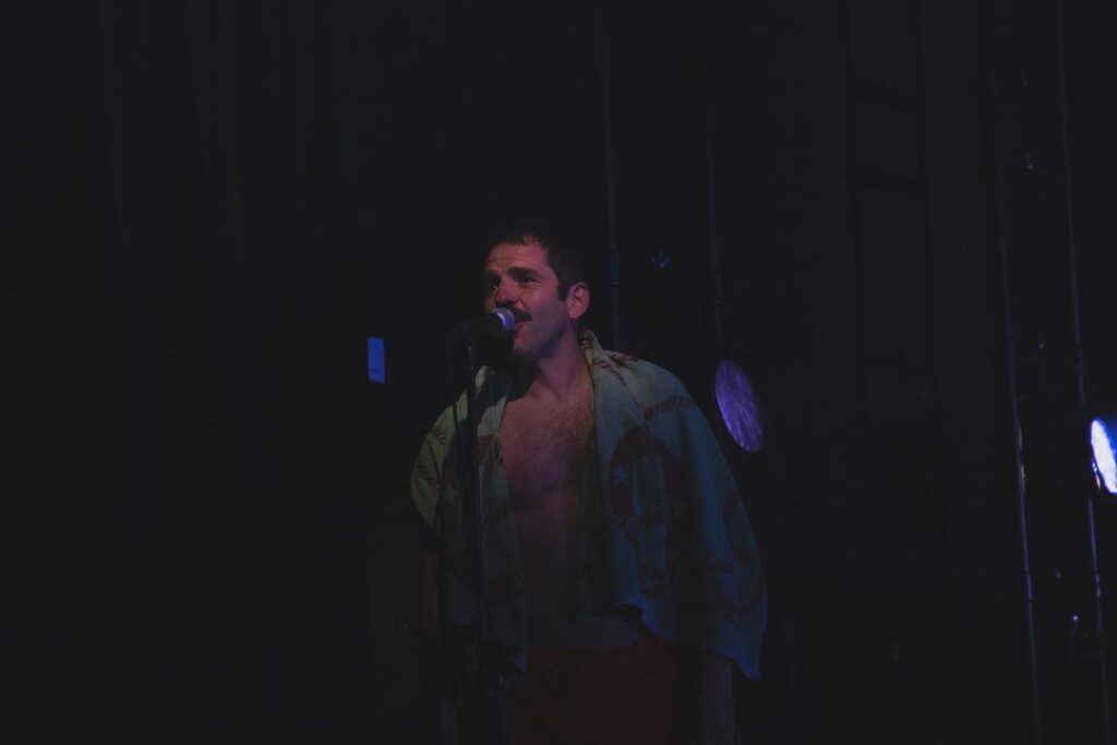 Un hombre está de pie en el escenario, bajo una luz tenue, hablando o cantando por un micrófono. Va sin camiseta y lleva una camisa estampada echada sobre los hombros; las luces del escenario proyectan un suave resplandor a sus espaldas.