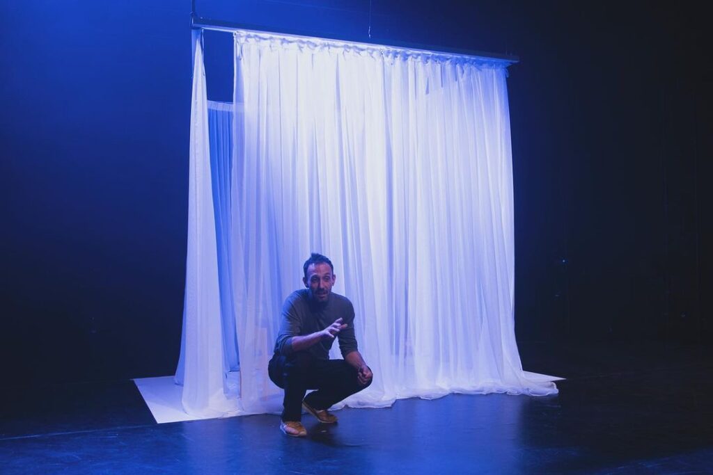 Una persona está agachada en un escenario oscuro, frente a un gran telón blanco y transparente que cuelga desde arriba, iluminado por luces azules.